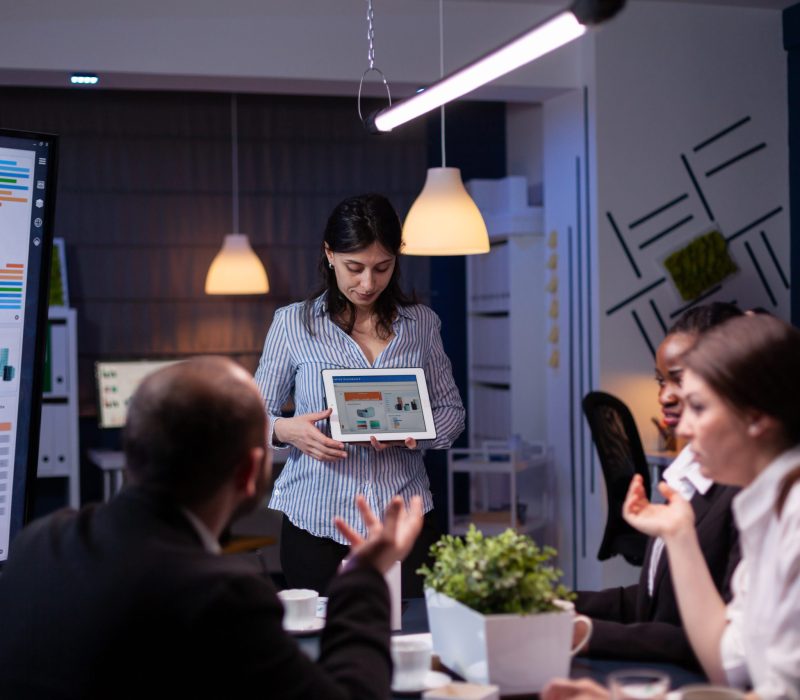 Overworked workaholic entrepreneur woman shwoing marketing graphs using tablet overworking at company solution late at night in meeting room. Diverse multi-ethnic teamwork solving financial strategy